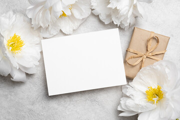 Blank wedding invitation mockup with copy space surrounded by white peonies and a gift box tied...