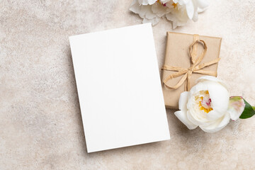 Blank greeting card mockup with copy space beside a wrapped gift and peony flowers on a textured...