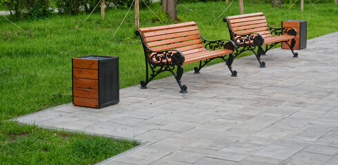 Two empty wooden benches with ornate iron frames and matching trash bins line a tiled pavement beside a green lawn in a summer city park, ideal for a quiet break. Photo. High resolution