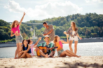 Group of cheerful young friends enjoying music and drinks outdoors in a scenic park on a sunny summer day
