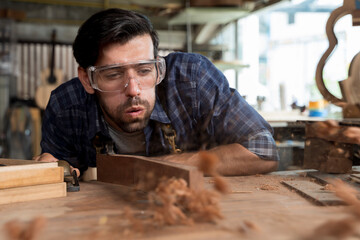 Carpenter blowing sawdust off wood surface while working in workshop, wearing safety goggles, close-up