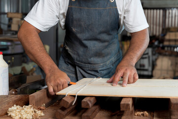 Close-up of craftsman’s hands cutting wood board with precision in workshop while wearing denim apron and using string
