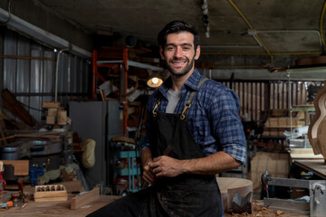 Confident carpenter standing in workshop smiling at camera while wearing apron and holding chisel with both hands