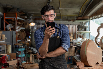 Young male carpenter in safety glasses using smartphone in wood workshop, modern craftsman lifestyle
