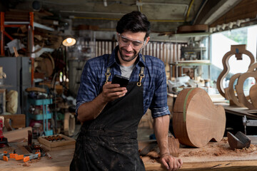 Smiling carpenter checking smartphone during break in workshop while wearing safety goggles and dirty apron