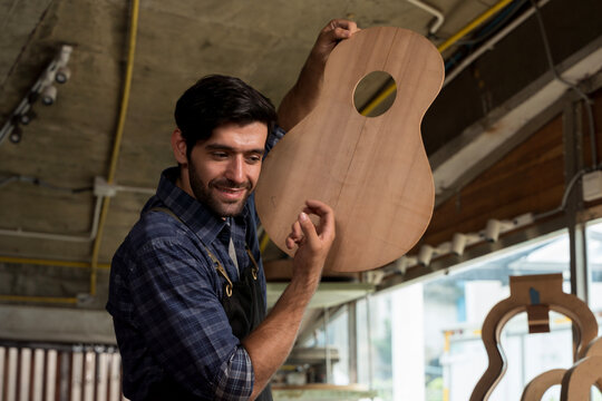 Craftsman smiling while holding unfinished wooden guitar body in workshop, showing detail of instrument build
