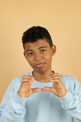 Vertical image of a dark skinned Latino teenager wearing a pastel blue sweatshirt making a broken heart with his hands on a light brown background with copy space, young man making a broken heart