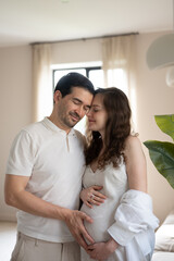Happy man and pregnant woman gently touching belly with hands, enjoying pregnancy time together in cozy bedroom