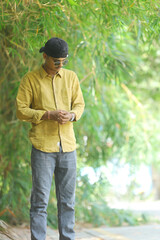 A stylish young man wearing a mustard yellow shirt, grey jeans, and sunglasses poses confidently in...