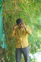 A stylish young man wearing a mustard yellow shirt, grey jeans, and sunglasses poses confidently in...
