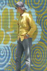 A stylish young man wearing a mustard yellow shirt, grey jeans, and sunglasses poses confidently in...