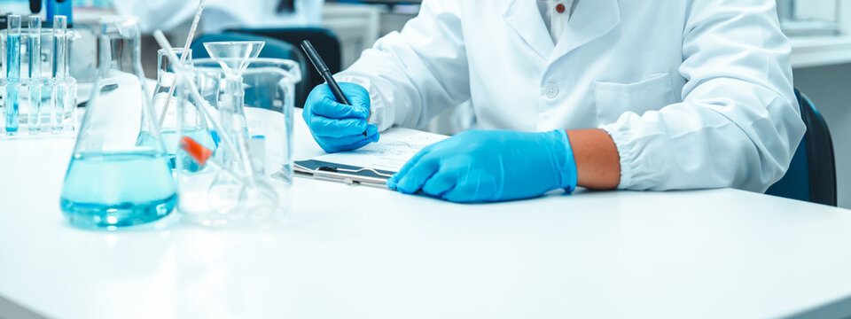 Young Asian male scientist recording data in a modern laboratory. Wearing safety goggles and gloves, surrounded by laboratory glassware. Biotechnology research and scientific documentation in progress