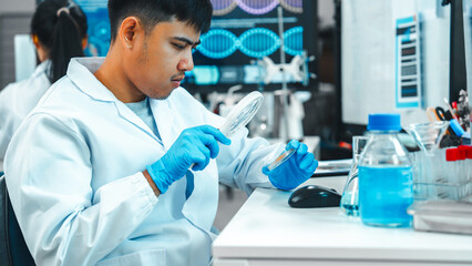 Young Asian scientist inspecting a petri dish with visible microbial growth using a magnifying...