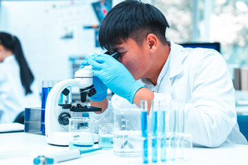 Scientist wearing lab gloves and coat using microscope in laboratory. Blue liquid in glassware...