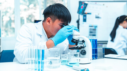 Scientist wearing lab gloves and coat using microscope in laboratory. Blue liquid in glassware...
