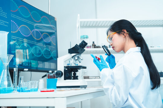 Female scientist in lab coat examining a petri dish with a magnifying glass in a modern laboratory. DNA structure displayed on screen. Concept of biotechnology, microbiology, and research.