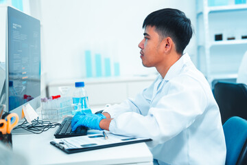 Male scientist in a lab coat and gloves working on a desktop computer in a modern laboratory. DNA charts and test tubes in the background. Concept of scientific research and data analysis.