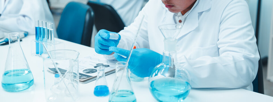 Asian male scientist wearing safety goggles and gloves working with blue liquid in a laboratory. Other researchers are working in the background in a clean, modern scientific environment. - Powered by Adobe