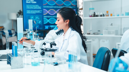 Fototapeta premium Asian female scientist in a white lab coat taking notes during a scientific experiment in a modern laboratory. Test tubes, microscope, and digital equipment are placed on the workbench.