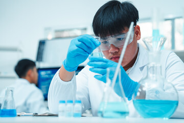 Asian male scientist wearing lab coat and safety goggles using pipette to transfer blue liquid in...