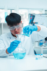 Fototapeta premium Male scientist in white lab coat and protective eyewear carefully pouring blue liquid into a glass flask using a funnel in a modern laboratory. Science experiment and chemical research in progress.