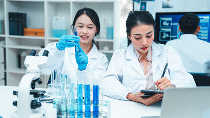 Two female scientists in lab coats working together in a science laboratory. One takes notes on a...