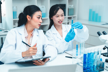 Two female scientists in lab coats working together in a science laboratory. One takes notes on a...