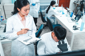 Male scientist examining a specimen under a microscope while a female scientist records...