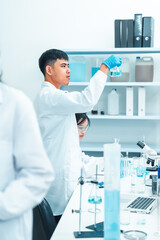 Fototapeta premium Male scientist holding a flask with blue liquid for examination in a laboratory. Other researchers work in the background. Scene reflects scientific research, analysis, and lab teamwork.