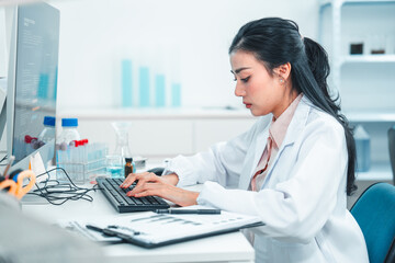 Female scientist in lab coat typing on a keyboard while analyzing research data on a computer. Laboratory equipment and chemical samples are visible, showing a scientific and focused work environment.