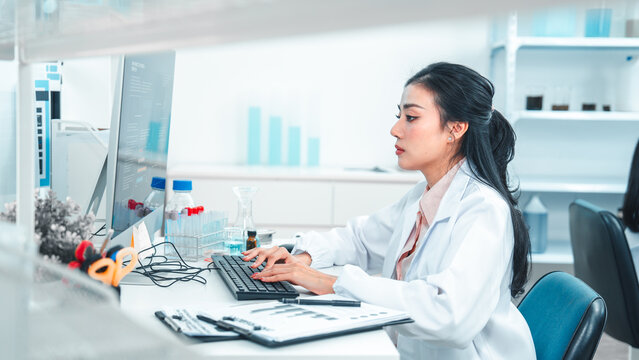 Female scientist in lab coat typing on a keyboard while analyzing research data on a computer. Laboratory equipment and chemical samples are visible, showing a scientific and focused work environment.
