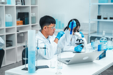 Fototapeta premium Two scientists in a laboratory working with test tubes. holds up a blue liquid sample while the woman observes attentively. Both wear lab coats, gloves, and safety goggles. Microscope in foreground.