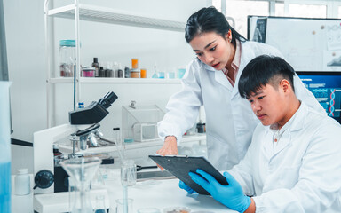 Female senior scientist supervising young male researcher in a modern lab. She reviews data on a...