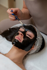 Beautician applying black mask treatment to woman's face in spa