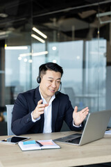 Vertical photo of a smiling young Asian male businessman in a headset sitting at his desk, talking...