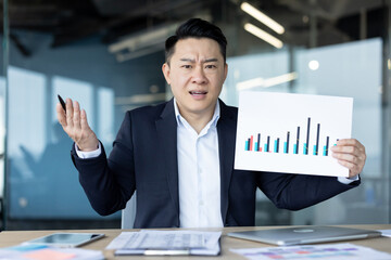 Portrait of a young serious Asian man sitting in the office at his desk, showing documents with...