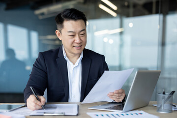 Asian smiling young male businessman sitting in the office at the desk in a suit and working with...