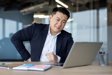 A young Asian man, twisted in pain, sits at his desk in the office and holds his back and lower...