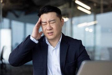 Asian young male businessman sitting in the office at his desk and holding his head with his hand...