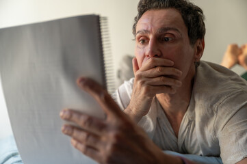 Shocked man reading a letter in bed covering his mouth with his hand