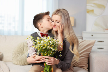 Little boy kissing his mother with bouquet of beautiful chamomile flowers on sofa at home
