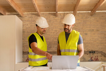 Two Construction workers in safety vests and helmets using laptop during house renovation project