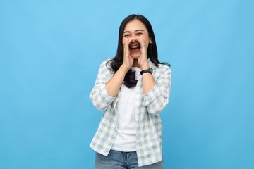 Fototapeta premium Young asian woman wearing casual clothes, shouting sharing news, giving announcement, advertising promotion or discount, looking at camera with hand near mouth, standing isolated on blue background.