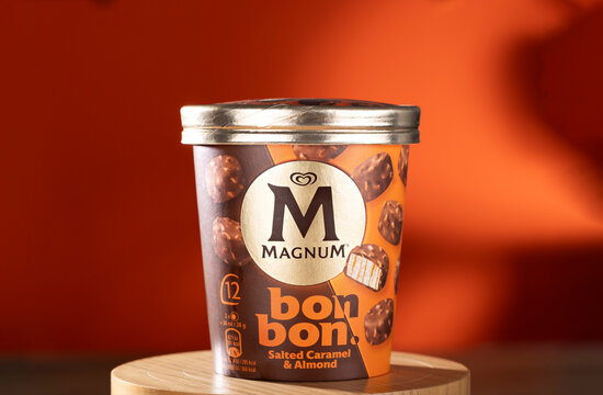 Studio shot of Magnum Bonbon Salted Caramel & Almond ice cream pack on a wooden stand, set against a vibrant orange background. Editorial image focusing on premium frozen dessert packaging.
