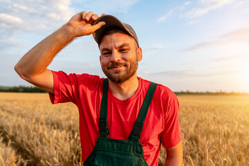 Portrait of cheerful man farm worker in cap and bib overalls with cereal field on background.