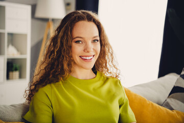 Smiling young woman with curly red hair wearing a green t-shirt sitting comfortably in a cozy...