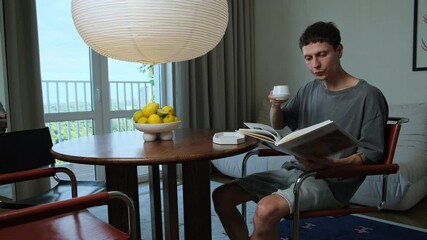 Young man enjoying morning coffee and reading a book at home, cozy interior with daylight and fresh lemons on the table.