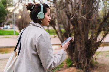 Young stylish influencer walking in a park listening to music with headphones and holding a smartphone