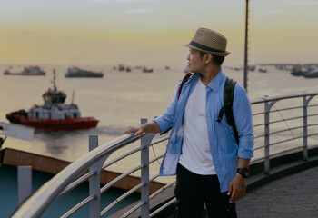 Young asian man in backpack and hat enjoying beautiful sunset solo traveling on the ship. Adventure and journey lifestyle concept.