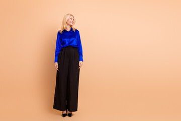 Elegant mature blonde woman in blue blouse and black pants on plain beige background with formal...